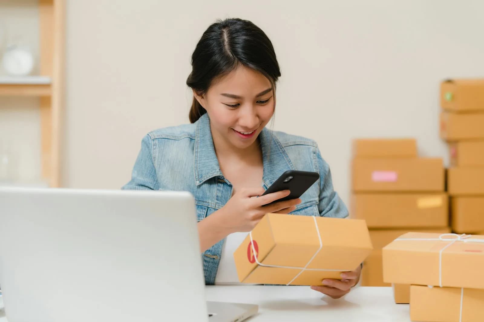 A small business owner checking a parcel and phone beside stacked shipping boxes